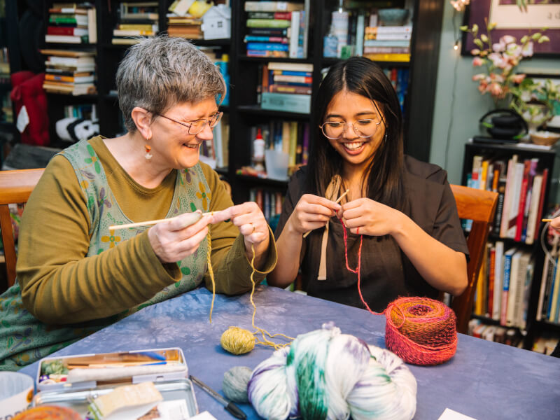 Two women smiling as they knit at a table together using yellow and red yarn, as well as knitting needles 